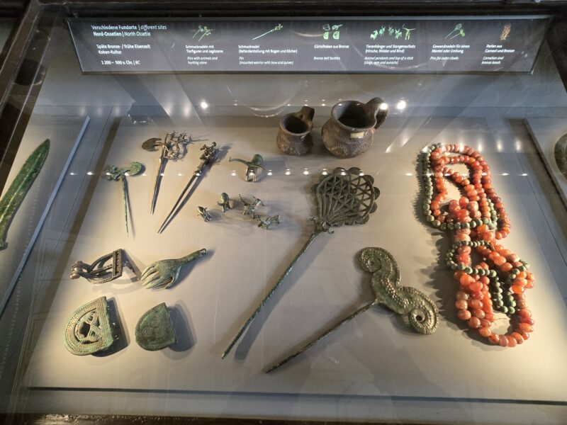 Bronze Age display: pins, vessels, and carved metal ornaments in a glass case, with orange bead necklace nearby.