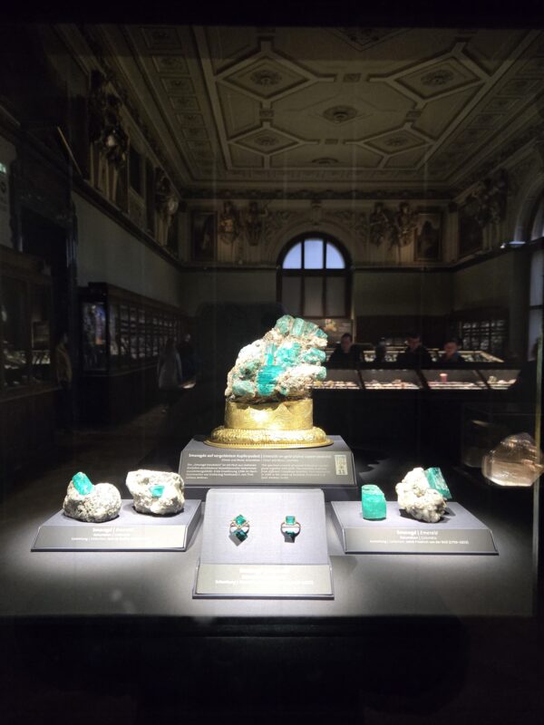 Display of emerald crystals and turquoise minerals on a gold pedestal inside a glass case at a museum exhibit. The ornate ceiling and gallery in the background set a historic viewing space.
