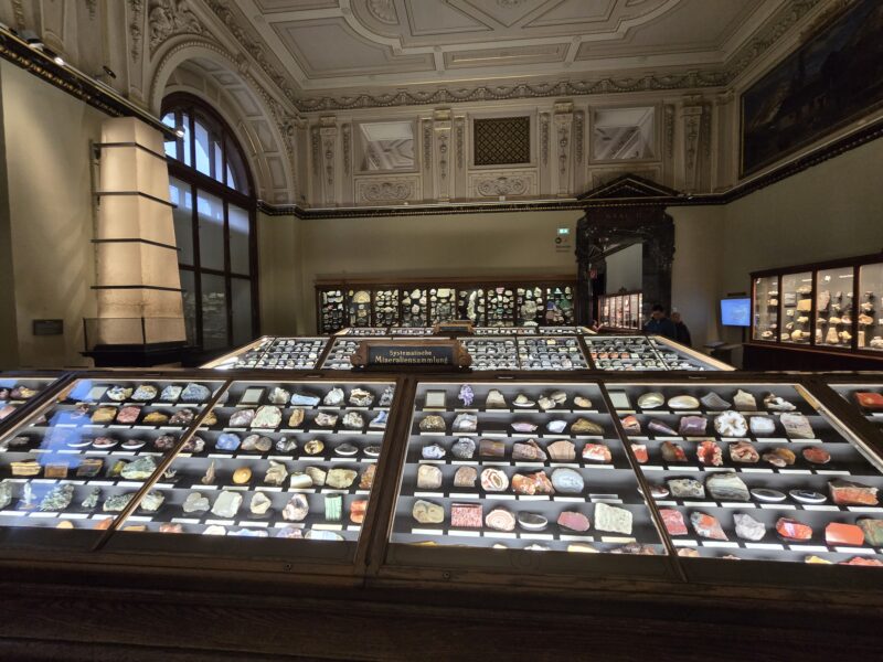 Ornate museum hall with glass display cases filled with mineral specimens and a sign reading 'Systematische Mineraliensammlung'