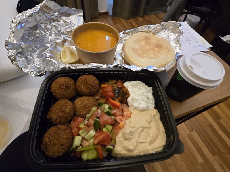 To-go meal on foil: falafel balls with hummus, chopped cucumber and tomato salad, pita bread, and a lemon wedge; cup of sauce on the side.