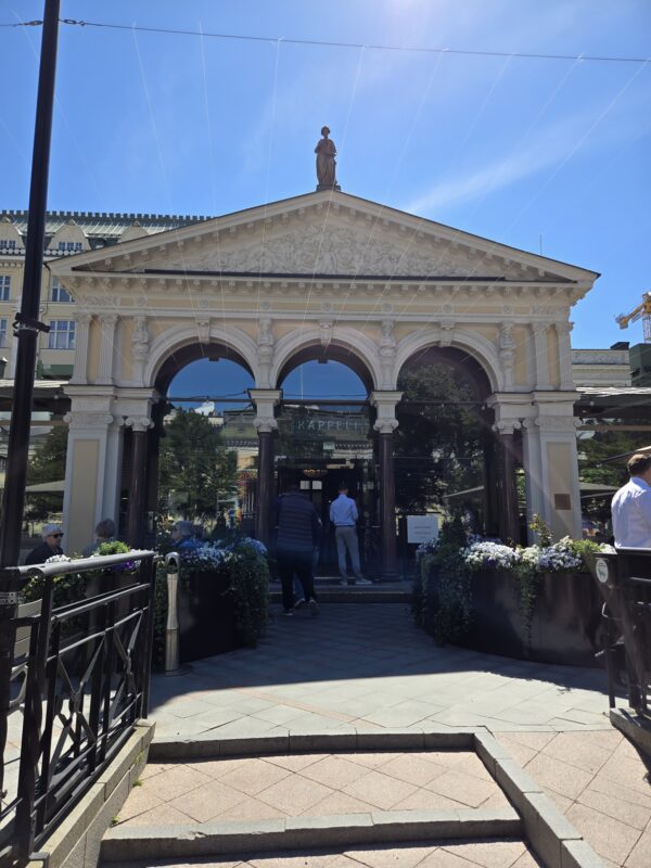 Elegant historic archway entrance with ornate carvings and a statue atop, blue sky overhead, people entering the building.